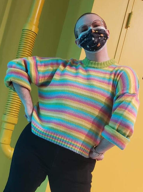 A photo of Orion, a trans person with a shaved head, glasses, and a mask with his hands on his hips, smiling.