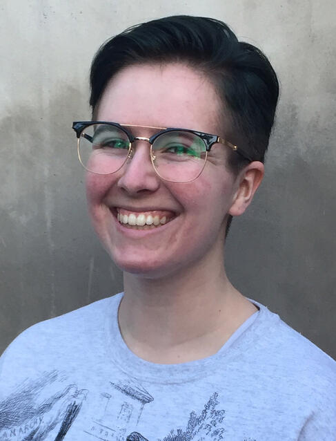 A photo of Tal, a white nonbinary person with short brown hair and glasses, smiling at the camera.